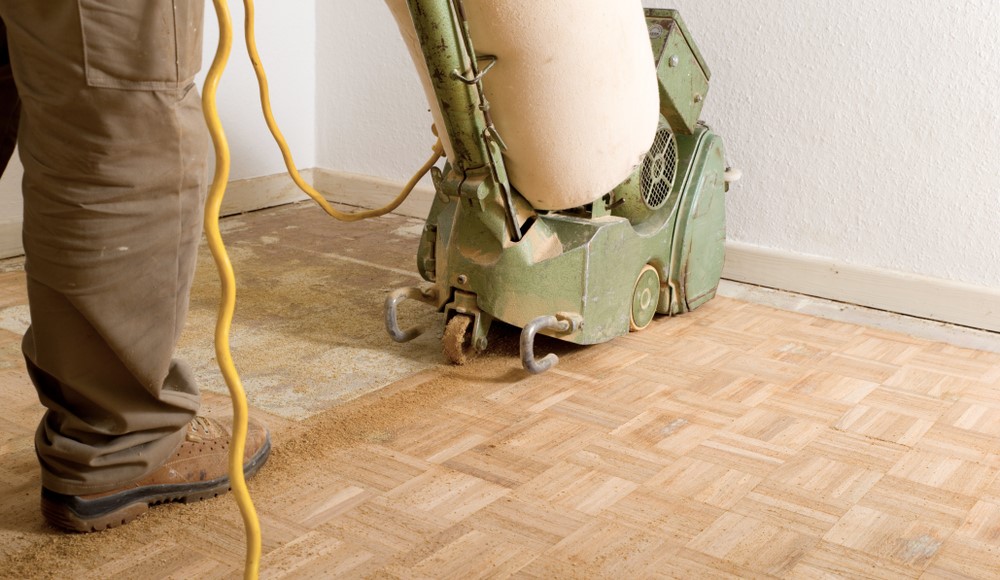 Wood Floor Sanding in Walthamstow: contractor guiding an industrial belt sander over a square-mosaic parquet hardwood floor, lifting old varnish and dust.