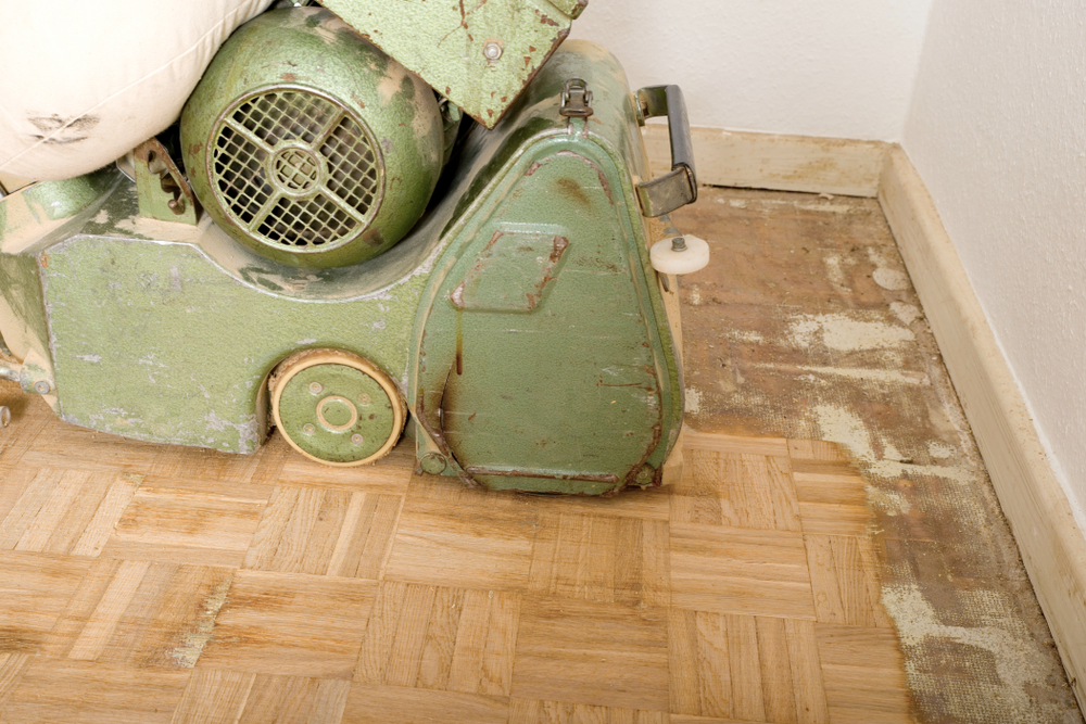 Wood Floor Sanding in Walthamstow – close-up of an industrial drum sander working along the edge of an oak parquet block floor, stripping away old adhesive.
